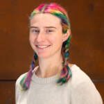 Headshot of a young woman. She has colourfully-dyed hair plaited into two braids, and wears a black longsleeve jumper.