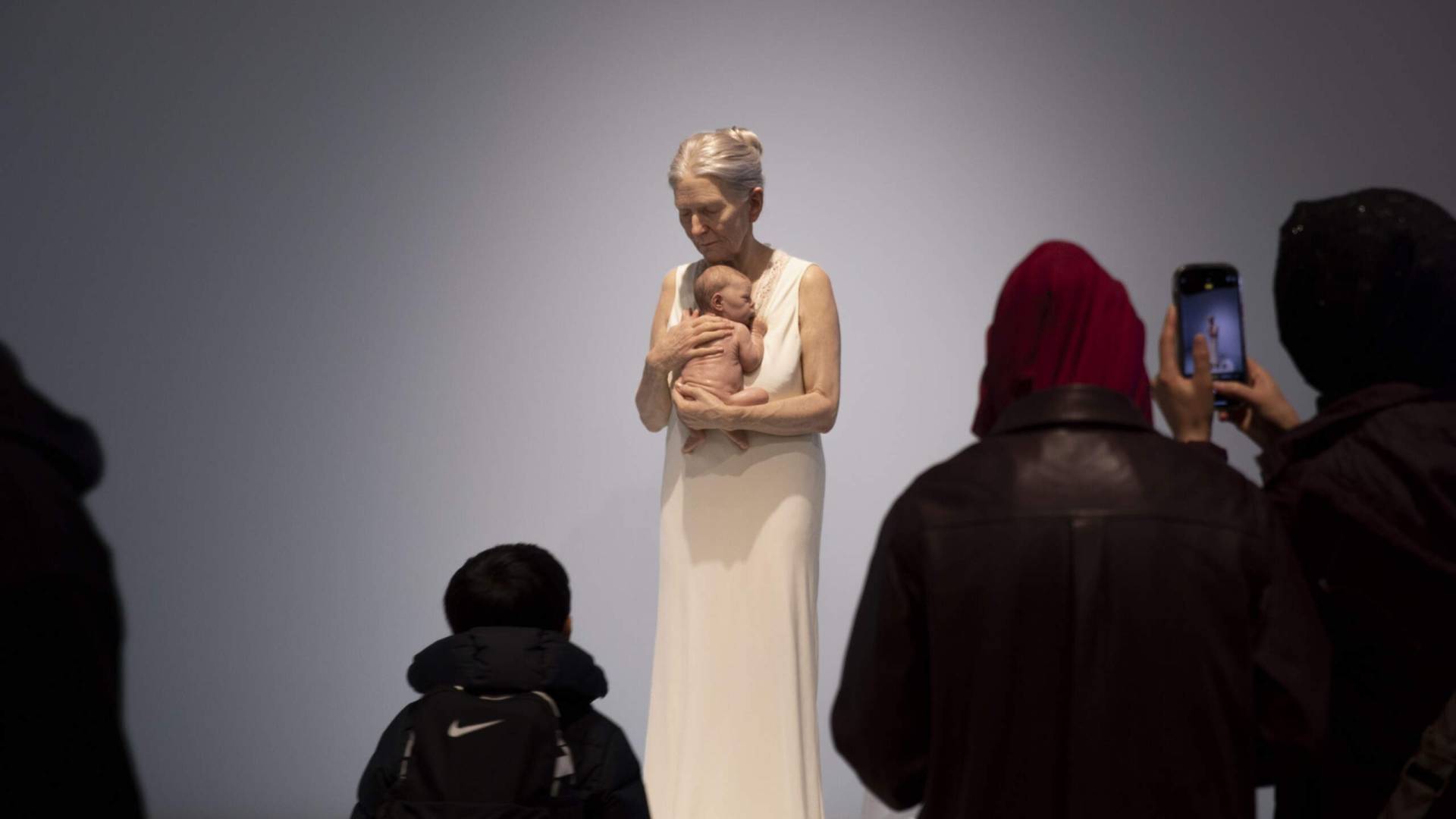 People view a hyperrealistic sculpture of an elderly woman holding a newborn baby in an art gallery
